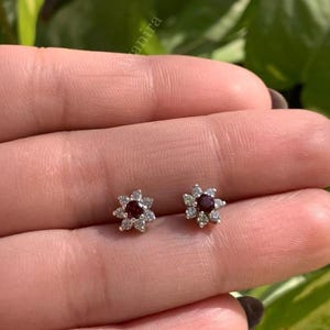 Może przedstawiać: Para małych kolczyków w kształcie kwiatów. Każdy kolczyk ma ciemnoczerwony kamień centralny otoczony małymi, przezroczystymi kamieniami akcentującymi. Kolczyki są osadzone w jasnym metalu, prawdopodobnie srebrze lub białym złocie.