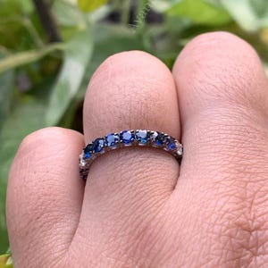 Pode incluir: Um anel de prata com uma faixa de pedras preciosas azuis e transparentes alternadas. O anel é usado em um dedo, com um fundo verde desfocado.