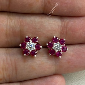 Puede incluir: Un par de pendientes de botón con una piedra preciosa central redonda y transparente rodeada por seis piedras redondas de color rojo intenso dispuestas en forma de flor, en metal plateado. La palabra Avanira es visible.
