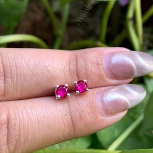 May include: A pair of small, square-cut, ruby-red gemstone stud earrings with a silver setting. The earrings are displayed on a person's finger, with green foliage in the background. The earrings are a classic jewelry item.