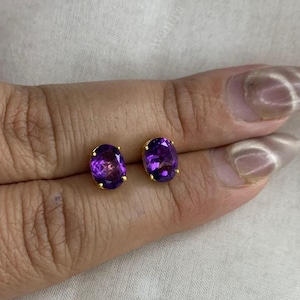 Può includere: Un paio di orecchini a lobo in oro, ciascuno con una pietra di ametista ovale. Le pietre viola sono incastonate in castoni d'oro, creando un look classico. Gli orecchini sono esposti su una superficie chiara. Le pietre misurano circa 1 cm.