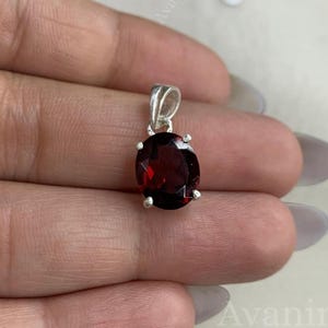 May include: A silver pendant with an oval, deep red gemstone. The gemstone is set in a silver bezel with four prongs. The pendant has a small silver loop at the top for a chain. The background is a light, neutral color.