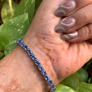 Puede incluir: Un brazalete plateado con piedras preciosas azules de forma ovalada. El brazalete se luce en una muñeca con un tatuaje de henna y uñas cuidadas. El fondo presenta follaje verde y el logotipo de Avanira.