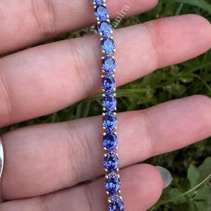 Puede incluir: Una pulsera de tenis que presenta una fila única de piedras preciosas de color azul violeta con corte ovalado, engastadas en una cadena de metal de tono plateado pulido. La pulsera muestra un patrón consistente de piedras facetadas.