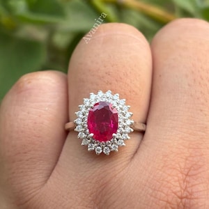 Può includere: Un anello color argento con una gemma rossa centrale dal taglio ovale, circondata da un doppio alone di piccole pietre chiare e scintillanti. La fascia in metallo è sottile e lucida, e la parola Avanira è visibile sullo sfondo.