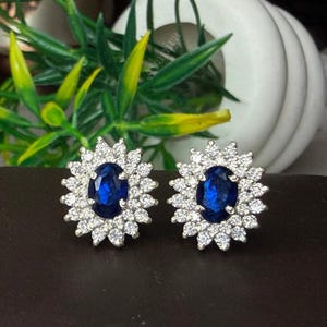 Pode incluir: Um par de brincos com pedras preciosas ovais azuis escuras, cada um rodeado por um halo de pedras menores e claras. Os brincos são definidos em metal prateado e são exibidos em uma superfície marrom escura.