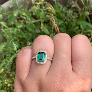 May include: A gold ring with a rectangular, emerald-cut green gemstone, encircled by a halo of smaller, clear gemstones. The ring's band is also set with small, clear gemstones. The ring is displayed against a backdrop of green foliage.