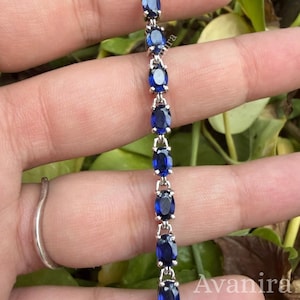 May include: A silver-toned tennis bracelet featuring a series of oval-cut, deep blue gemstones set in a linked metal chain. The bracelet is displayed vertically, showcasing the uniform arrangement of the blue stones held by small metal prongs.