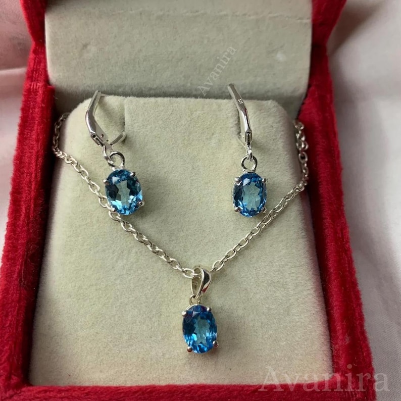 May include: A jewelry set featuring a silver chain necklace with a blue oval pendant and matching blue oval earrings. The set is displayed in a red velvet jewelry box. The earrings have a hook closure.