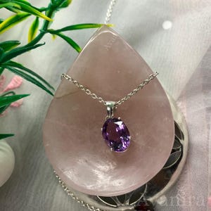 Puede incluir: Un collar de plata con un colgante de piedra preciosa ovalada p&uacute;rpura. El colgante est&aacute; engastado en un engaste de plata y cuelga de una delicada cadena de plata. El collar se exhibe sobre una gran piedra rosa en forma de l&aacute;grima.