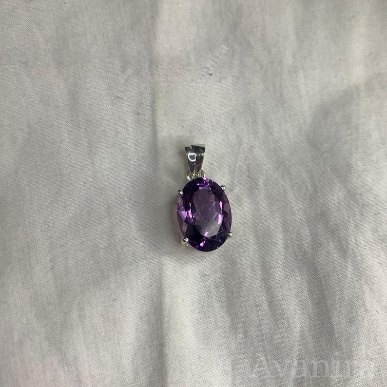 Puede incluir: Un colgante de plata con una gran amatista ovalada. La piedra de color p&uacute;rpura oscuro est&aacute; engastada en un bisel de plata y tiene un peque&ntilde;o lazo para una cadena. El colgante se muestra sobre un fondo de tela blanca.