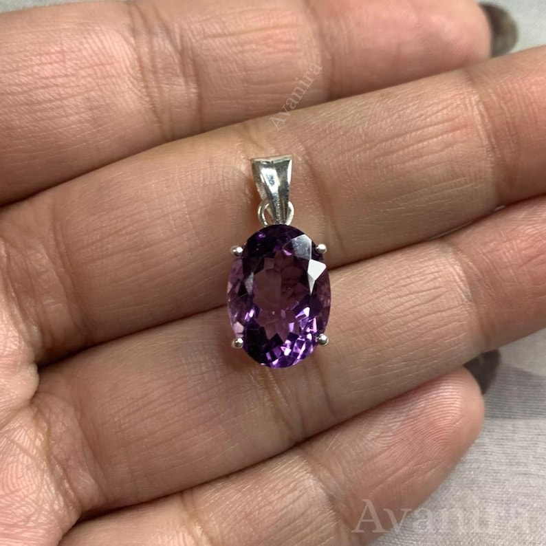 Puede incluir: Un colgante de plata con una piedra preciosa de amatista ovalada. La piedra de color p&uacute;rpura oscuro est&aacute; engastada en un engaste de cuatro puntas con una peque&ntilde;a argolla. El colgante se sostiene en una mano. La piedra mide unos 2 cm de largo.
