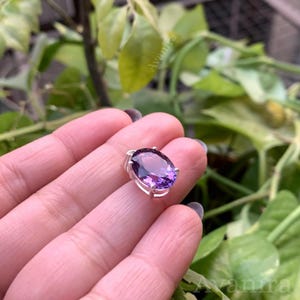 Puede incluir: Un colgante de plata con una piedra preciosa de amatista ovalada de color p&uacute;rpura intenso. La piedra preciosa est&aacute; engastada en un engaste de cuatro puntas. El colgante se sostiene en una mano sobre un fondo de follaje verde.