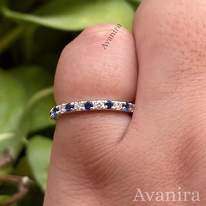 Può includere: Un anello a fascia color argento caratterizzato da un motivo alternato di pietre rotonde blu scuro e pietre bianche trasparenti incastonate lungo l'intera circonferenza. La parola Avanira è stampata in piccolo testo nero sulla fascia.