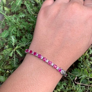 Op de afbeelding: Een zilveren armband met ovale, levendige rode edelstenen en kleinere, heldere stenen. De armband wordt getoond tegen groene bladeren. Het ontwerp is elegant.