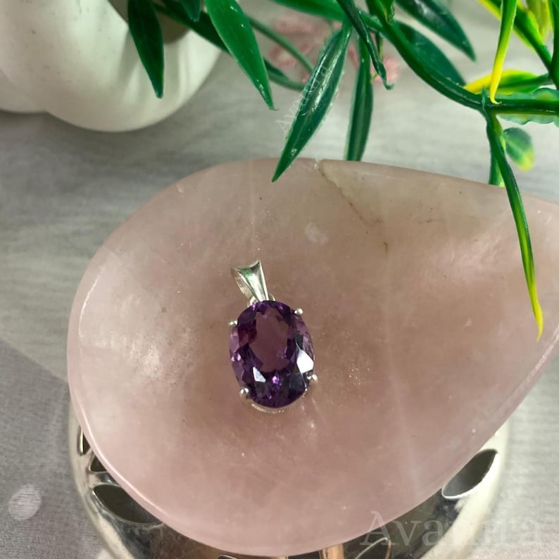 Puede incluir: Un colgante de plata con una piedra preciosa ovalada de color p&uacute;rpura oscuro. El colgante est&aacute; sobre una superficie rosa claro y redondeada, con follaje verde en el fondo. La piedra preciosa est&aacute; facetada.