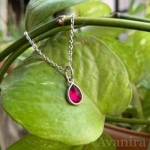 May include: A silver pendant necklace with a teardrop-shaped, deep red gemstone. The necklace hangs against a backdrop of vibrant green leaves, creating a striking contrast. The pendant is approximately 0.75 inches in length.