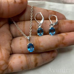 May include: A silver necklace with a blue oval pendant and matching silver earrings with blue oval gemstones. The jewelry set is displayed on a light background.