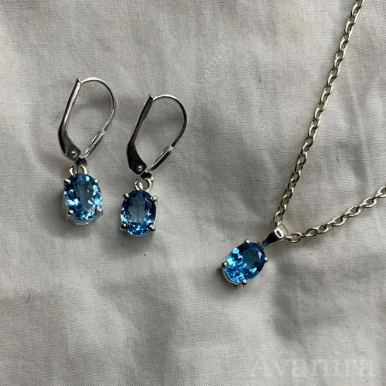 May include: A jewelry set featuring a pair of lever-back earrings and a necklace. Each piece has an oval, light blue gemstone set in a silver-colored metal. The necklace has a delicate chain.