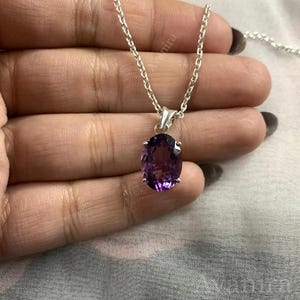 Puede incluir: Un collar de plata con un colgante de amatista ovalada de color p&uacute;rpura intenso. La piedra preciosa est&aacute; engastada en un bisel de plata y cuelga de una delicada cadena de plata. El collar se muestra sobre un fondo neutro.