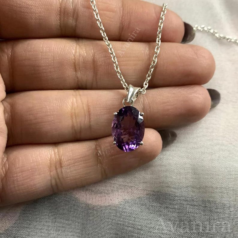 Puede incluir: Un collar de plata con un colgante de amatista ovalada de color p&uacute;rpura intenso. La piedra preciosa est&aacute; engastada en un bisel de plata y cuelga de una delicada cadena de plata. El collar se muestra sobre un fondo neutro.