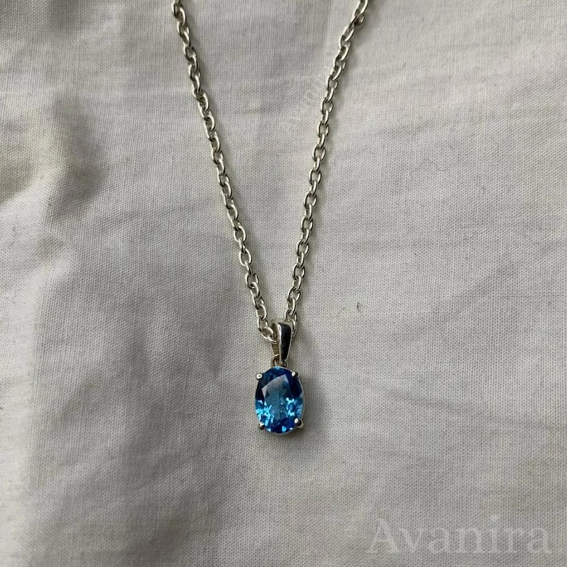 May include: A silver necklace with a blue oval gemstone pendant. The pendant is a vibrant shade of blue, set in a silver bezel. The chain is a delicate link style, and the necklace is displayed against a white background.
