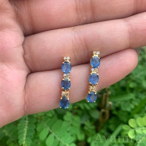 May include: A pair of gold earrings featuring three oval, blue gemstones and small, clear accent stones. The earrings are presented against a green backdrop.