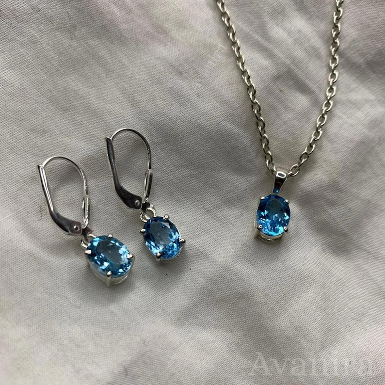 May include: A jewelry set featuring a silver necklace with a pendant and a pair of earrings. Each piece showcases a vibrant, oval-shaped, light blue gemstone. The set is displayed on a white, textured surface.