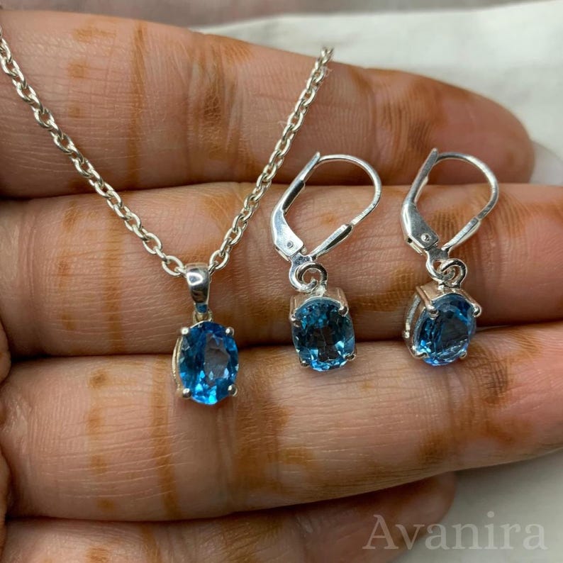 May include: A silver necklace with a blue oval pendant and matching leverback earrings. The jewelry set features a bright blue gemstone. The chain and earring hooks are silver. The jewelry is displayed on a hand.