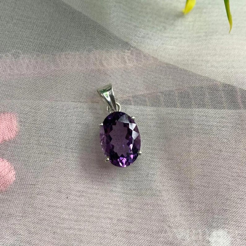 Puede incluir: Un colgante de plata con una piedra de amatista de talla ovalada. La piedra de color p&uacute;rpura oscuro est&aacute; engastada en un bisel de plata y tiene una superficie facetada. El colgante cuelga de una peque&ntilde;a fianza de plata.