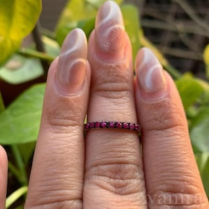 May include: A thin, silver ring set with small, round, red gemstones. The ring is worn on a finger, with the gemstones encircling the band. The ring is a piece of jewelry, likely an eternity ring or a similar style.