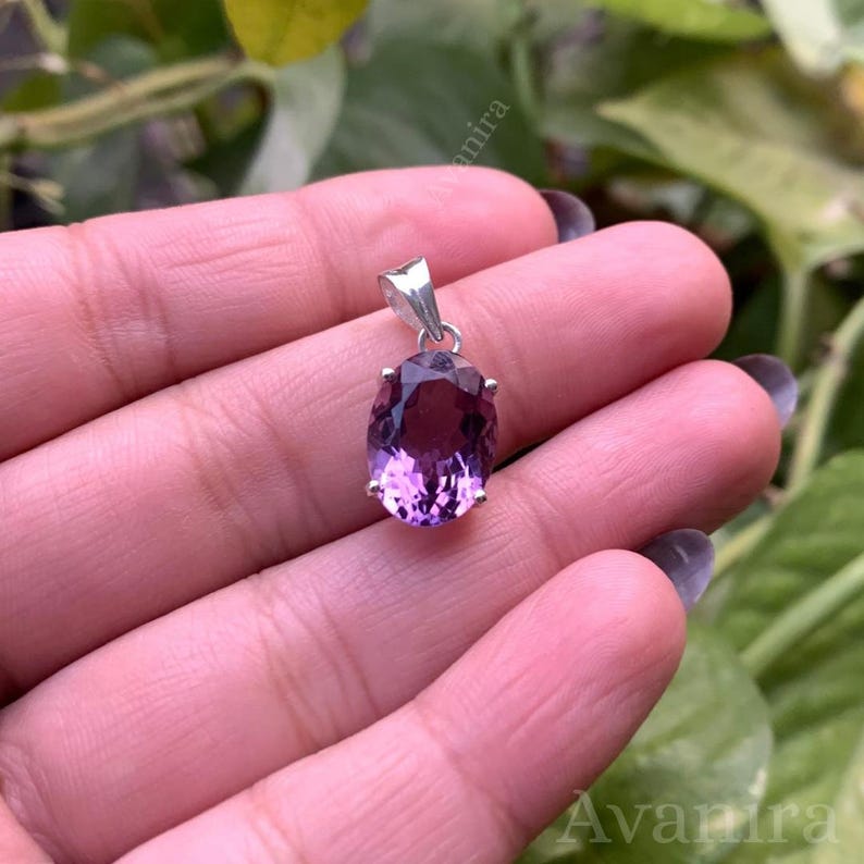 Puede incluir: Un colgante de plata con una amatista ovalada de color p&uacute;rpura oscuro. La piedra preciosa est&aacute; engastada en un bisel de plata y tiene un acabado pulido. El colgante est&aacute; suspendido de una peque&ntilde;a fianza de plata. Fondo borroso, con follaje verde.