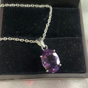 Puede incluir: Un collar de plata con un colgante de amatista ovalada. La gema p&uacute;rpura est&aacute; engastada en un engaste de cuatro garras y cuelga de una delicada cadena. El collar se exhibe en una caja de joyer&iacute;a negra.