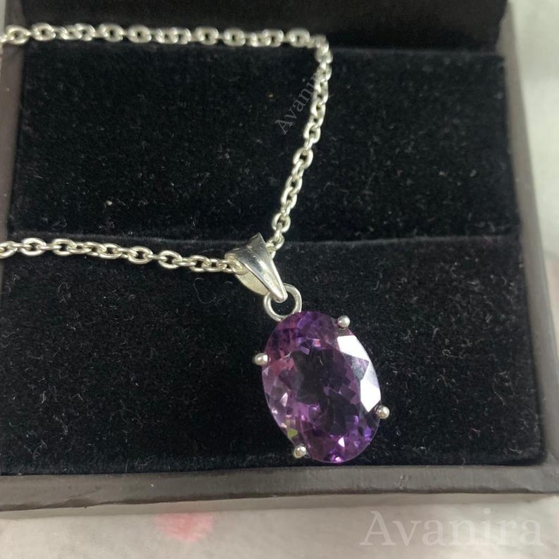 Puede incluir: Un collar de plata con un colgante de amatista ovalada. La gema p&uacute;rpura est&aacute; engastada en un engaste de cuatro garras y cuelga de una delicada cadena. El collar se exhibe en una caja de joyer&iacute;a negra.