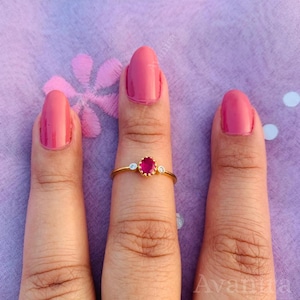 Puede incluir: Un anillo de oro con un rubí central ovalado y dos piedras preciosas claras más pequeñas. El anillo se muestra sobre un fondo morado. El diseño del anillo es sencillo y elegante.