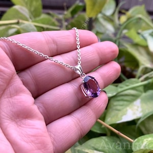 Puede incluir: Un collar de plata con un colgante de amatista ovalada. La gema p&uacute;rpura est&aacute; engastada en un bisel de plata y cuelga de una delicada cadena de plata. El fondo es un follaje verde borroso.