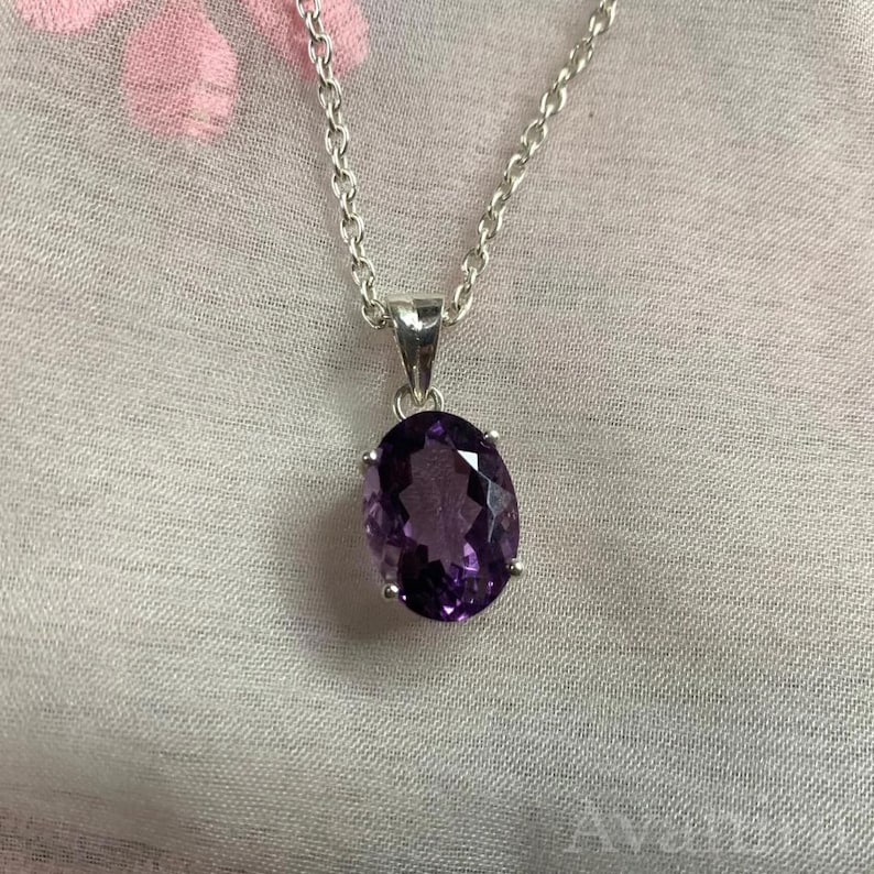 Puede incluir: Un collar de plata con un colgante de amatista ovalado. La gema de color p&uacute;rpura oscuro est&aacute; engastada en un bisel de plata y cuelga de una delicada cadena de plata. El fondo es una tela suave estampada.