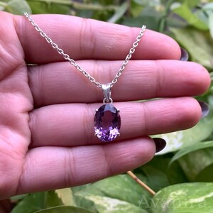 Puede incluir: Un collar de plata con un colgante con una gran gema de amatista de talla ovalada. La amatista muestra tonos de p&uacute;rpura y est&aacute; engastada en un bisel de plata. La cadena es delicada y parece ser de plata.