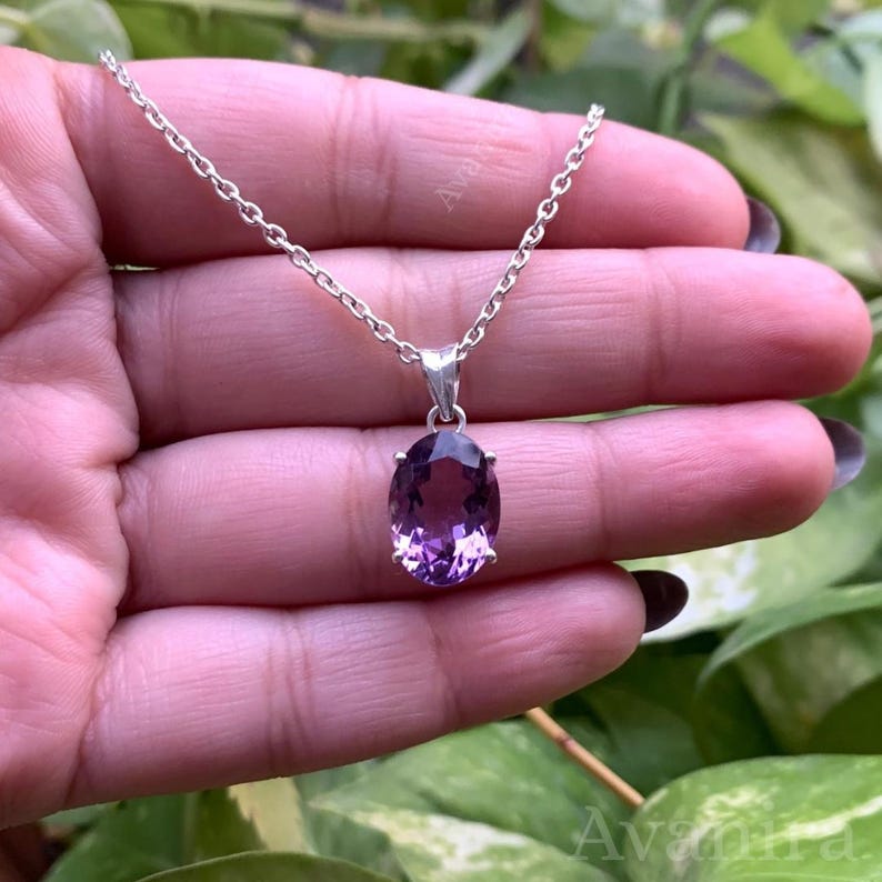 Puede incluir: Un collar de plata con un colgante con una gran gema de amatista de talla ovalada. La amatista muestra tonos de p&uacute;rpura y est&aacute; engastada en un bisel de plata. La cadena es delicada y parece ser de plata.