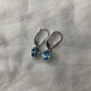 May include: A pair of silver leverback earrings, each featuring a light blue oval gemstone. The earrings are displayed on a white, textured surface. The gemstones have a slight sparkle.