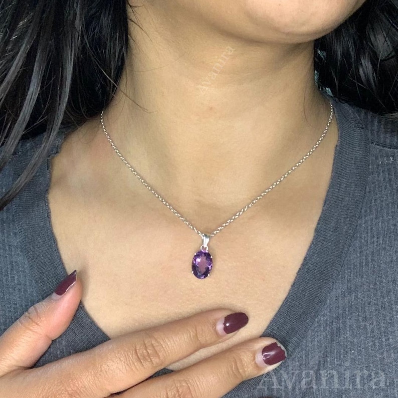 Puede incluir: Un collar de plata con un peque&ntilde;o colgante ovalado de amatista p&uacute;rpura. El colgante cuelga de una delicada cadena de plata. El collar se lleva alrededor del cuello de una persona.