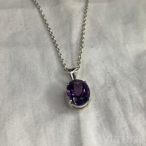 Puede incluir: Un collar de plata con un gran colgante ovalado de amatista de color p&uacute;rpura intenso. La piedra preciosa est&aacute; engastada en un bisel de plata y cuelga de una delicada cadena de plata. El fondo es una tela blanca texturizada.