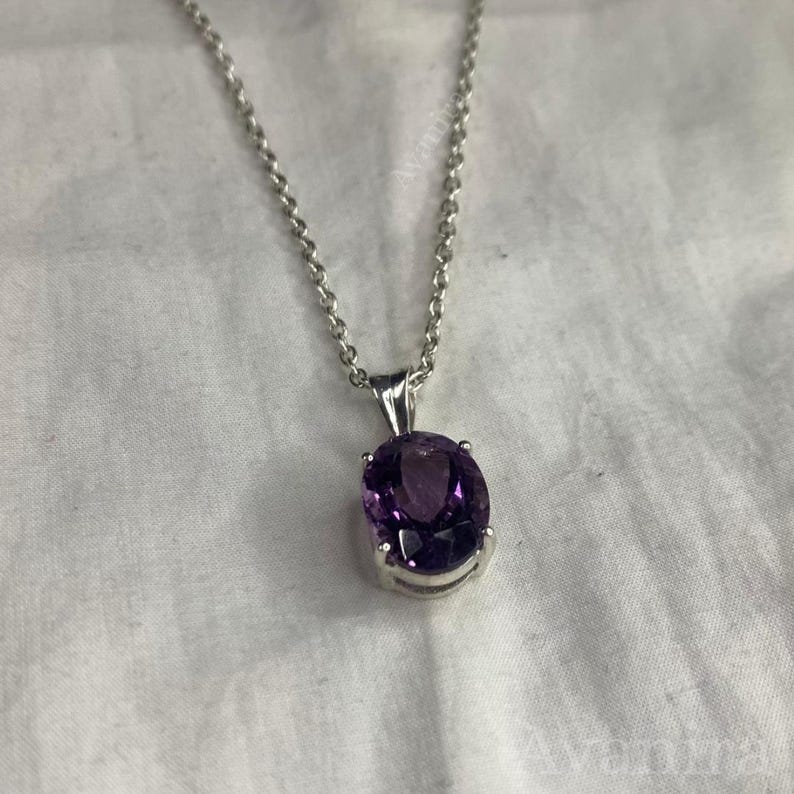 Puede incluir: Un collar de plata con un gran colgante ovalado de amatista de color p&uacute;rpura intenso. La piedra preciosa est&aacute; engastada en un bisel de plata y cuelga de una delicada cadena de plata. El fondo es una tela blanca texturizada.