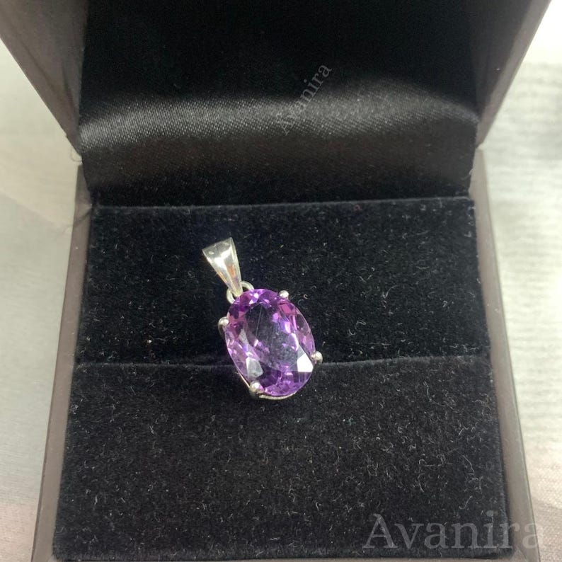 Puede incluir: Un colgante de amatista p&uacute;rpura con una fianza plateada, engastado en una caja de joyer&iacute;a de terciopelo negro. La gema de forma ovalada es de un tono p&uacute;rpura vibrante. El colgante se exhibe en un entorno oscuro y elegante.