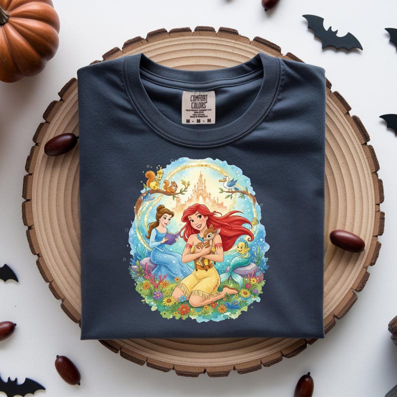 May include: Dark blue t-shirt featuring a colorful illustration of two princesses with a castle, animals, and flowers. The shirt has a crew neck and the Comfort Colors brand label.