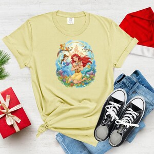 May include: Light yellow t-shirt featuring a colorful illustration of two princesses and a small animal within a circular frame. The princesses are depicted in a floral setting with a castle in the background. The shirt is paired with blue jeans and black sneakers.