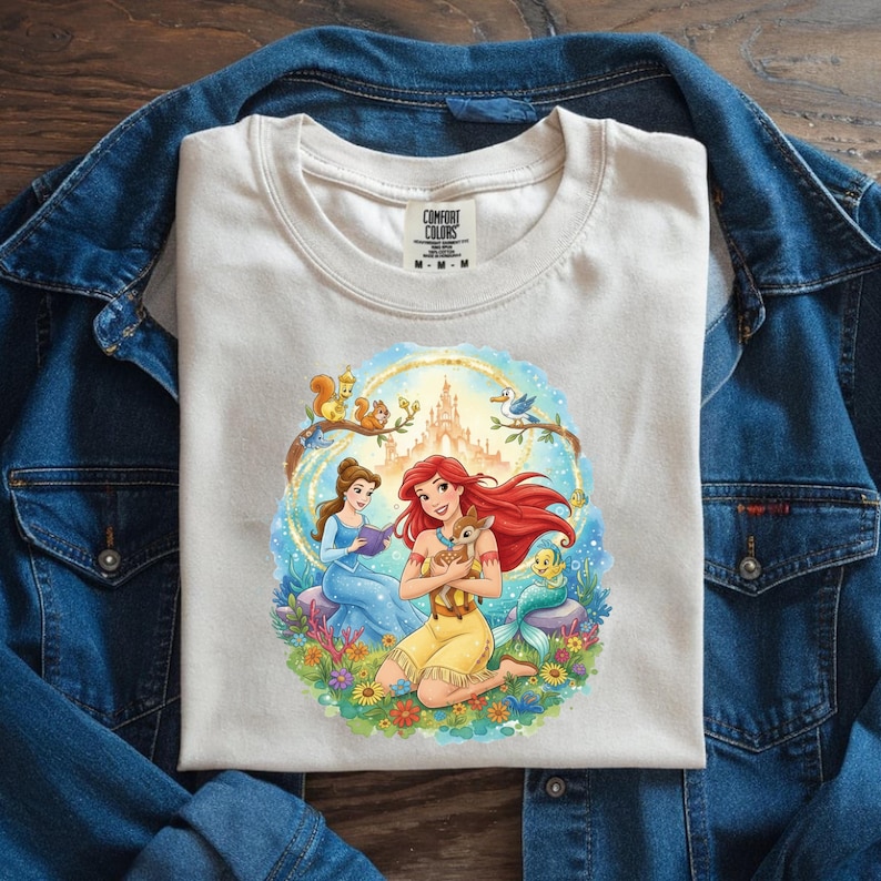 May include: Cream-colored t-shirt featuring a colorful illustration of Disney princesses Ariel and Belle, surrounded by woodland creatures and a castle. The shirt has the Comfort Colors logo on the collar and is laid on a denim jacket.