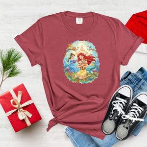 May include: A maroon t-shirt featuring a colorful graphic of two princesses with a castle in the background. The princesses are surrounded by flowers and animals. The shirt is styled with blue jeans and black sneakers.