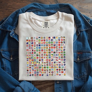 May include: A cream-colored t-shirt featuring a grid of colorful, circular flags from various countries. The shirt is labeled "Comfort Colors" and is displayed on a denim jacket. The flags are arranged in a neat pattern, showcasing a diverse range of national symbols.