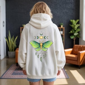 May include: Cream-colored hoodie featuring a large graphic of a green and yellow moth with moon phases and feathers. The moth design is centered on the back of the hoodie. The hoodie has a drawstring hood and long sleeves.